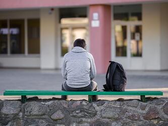 young man sits on bench, facing away from camera, with rucksack next to him.